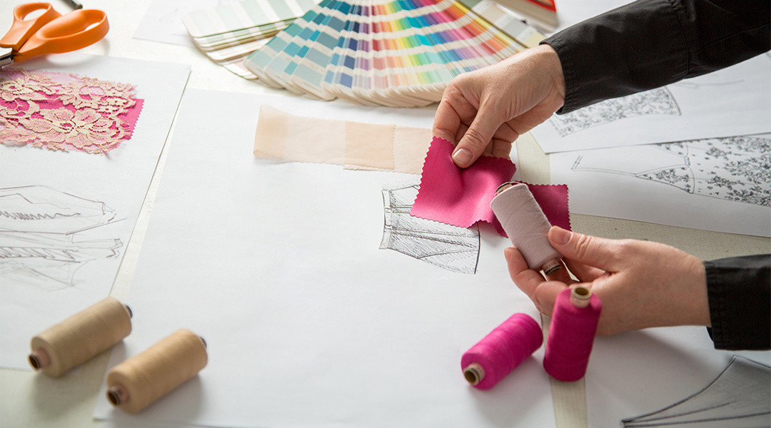 a designer's hands holding fabric swatches and thread over a fashion illustration